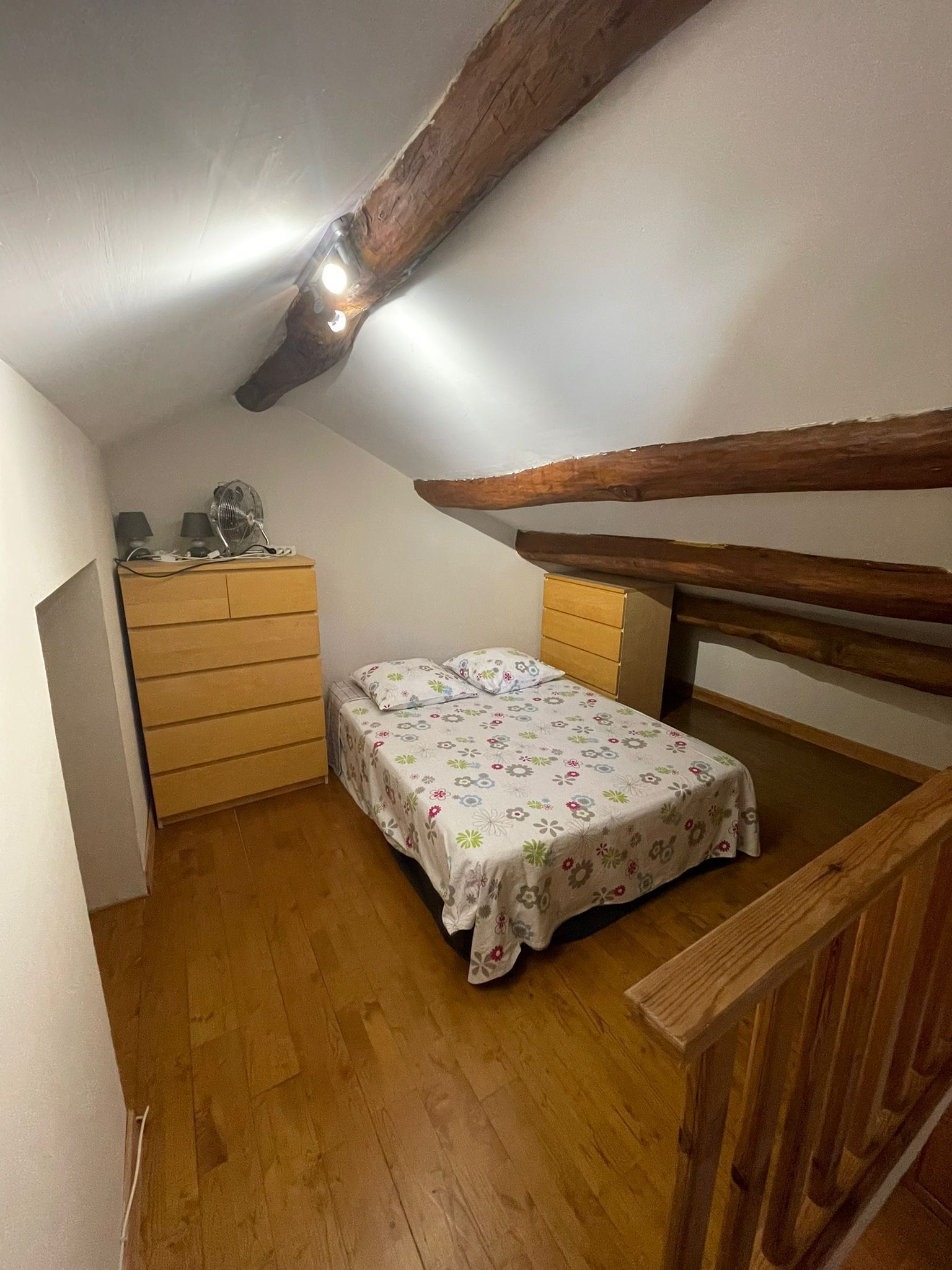 Un dormitorio cálido y acogedor en el ático, con una viga de madera en el techo, una cama individual con ropa de cama floral, un aparador y una mesita de noche con lámparas.