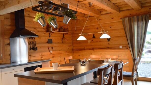 A wooden cabin interior with a kitchen area featuring a large stove, hanging pots, and a dining table with chairs.