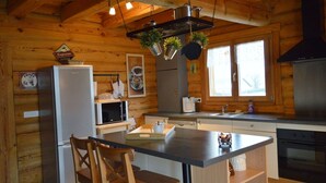 A wooden cabin kitchen with a central island, wooden chairs, a refrigerator, a microwave, a sink, and hanging plants.