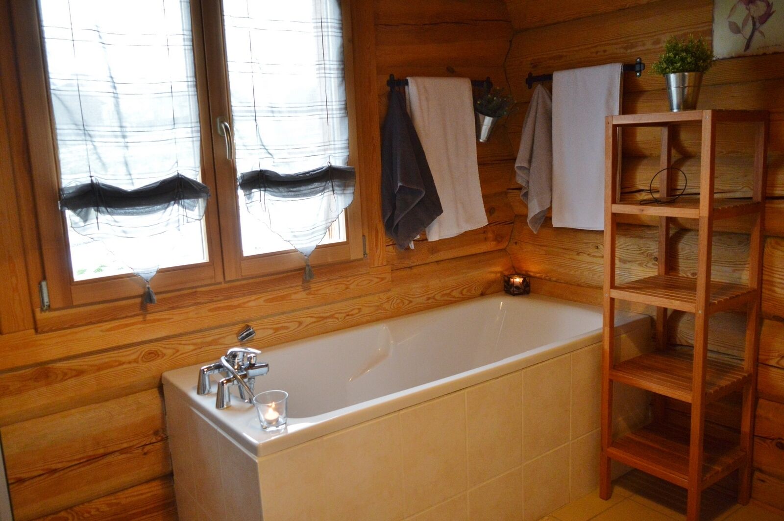 A wooden cabin bathroom with a bathtub, a window with curtains, a shelf with towels, and a small candle.