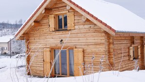 A wooden log cabin with a pitched roof, large windows with shutters, and a visible chimney.