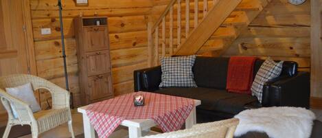 Interior acogedor de una cabaña de troncos con una escalera, muebles de mimbre, un mantel a cuadros y una chimenea.