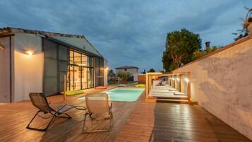 Un espace piscine extérieur moderne, doté d’une terrasse en bois, de chaises longues et d’un patio bien éclairé.
