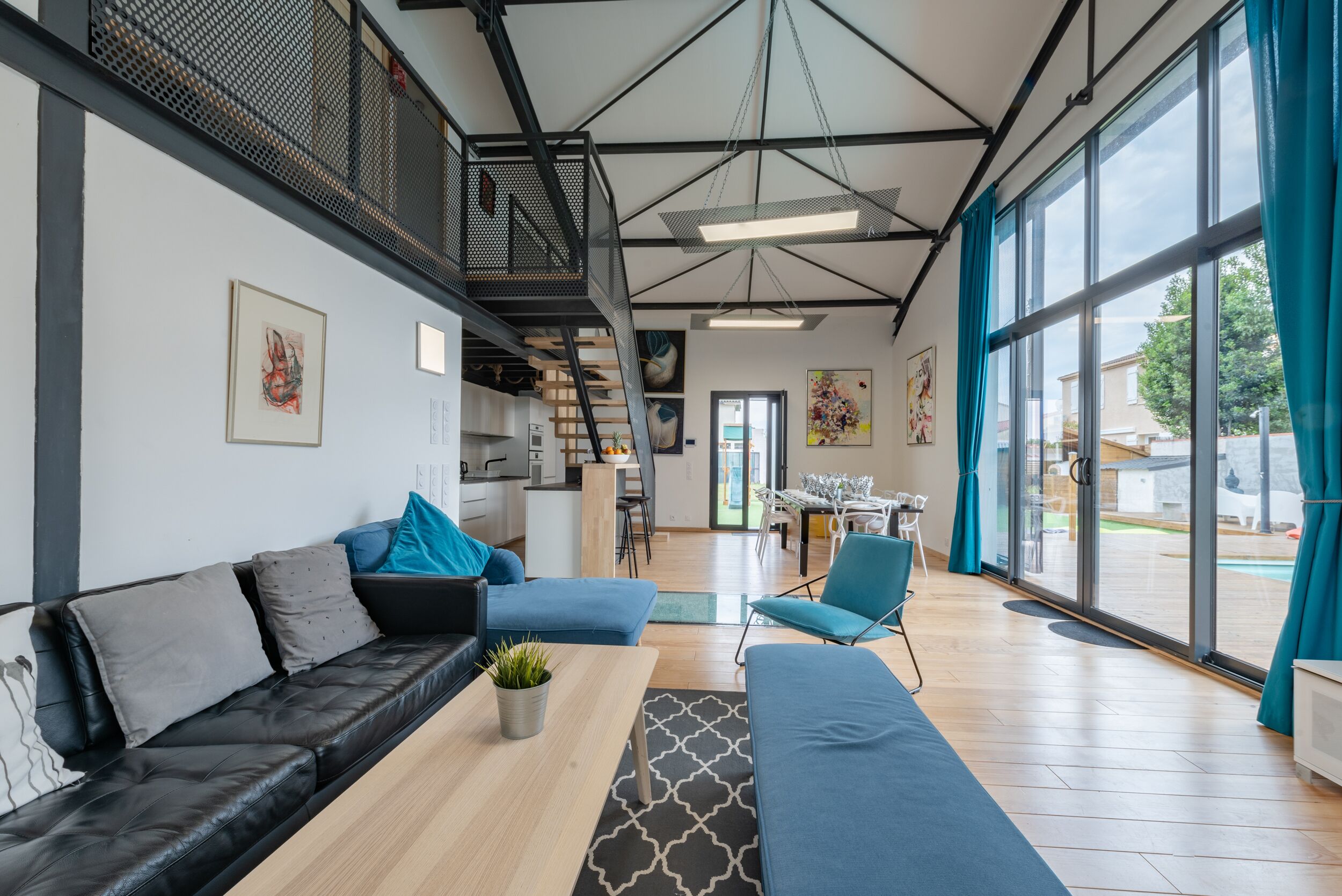 Un salon moderne avec un canapé en cuir noir, une table basse en bois et un fauteuil bleu. Un escalier avec une rampe en métal mène à l’étage supérieur.