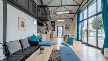 Un salon moderne avec un canapé en cuir noir, une table basse en bois et un fauteuil bleu. Un escalier avec une rampe en métal mène à l’étage supérieur.