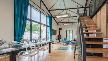 Une salle à manger spacieuse avec une longue table, des chaises blanches et des rideaux turquoise. Un escalier avec des marches en bois et une rampe en métal mène à un niveau supérieur.