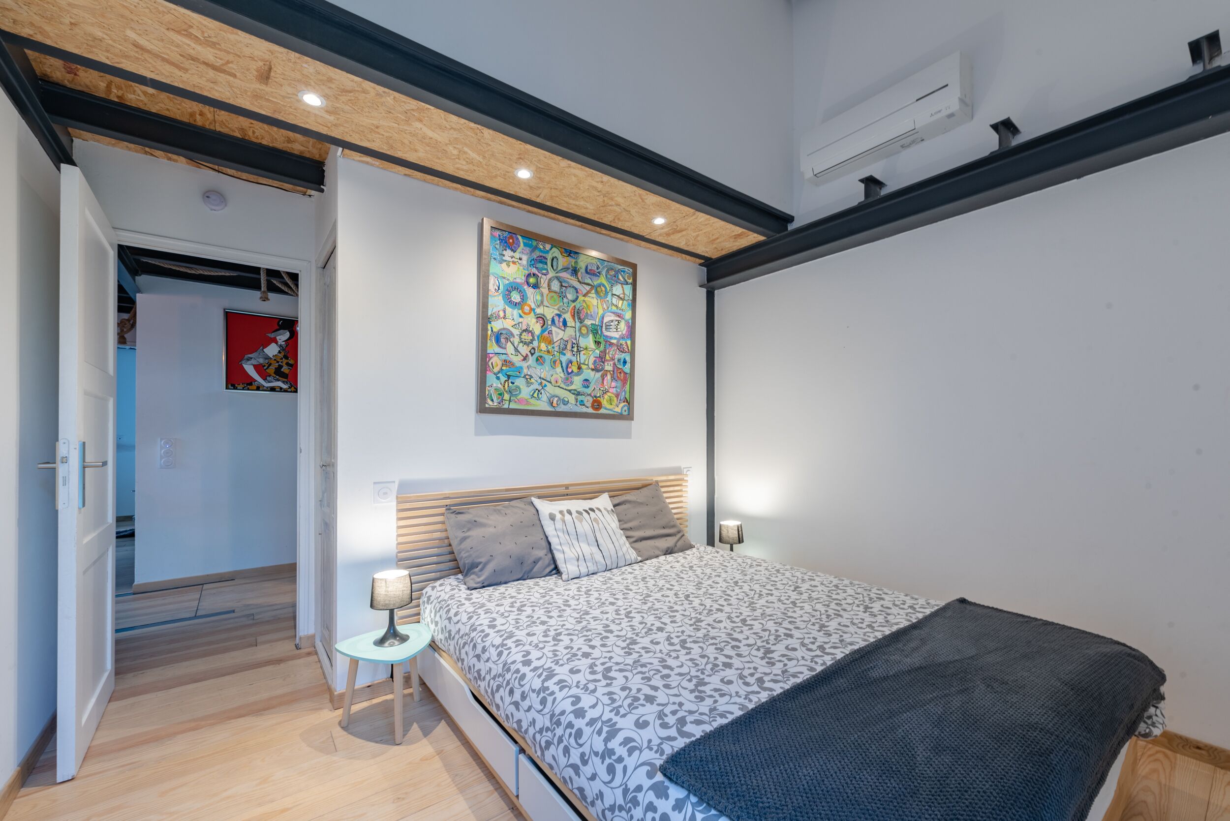 Une chambre moderne avec un plafond en bois, un grand lit avec une literie à motifs, un tableau abstrait aux couleurs vives et une petite table de chevet avec une lampe.