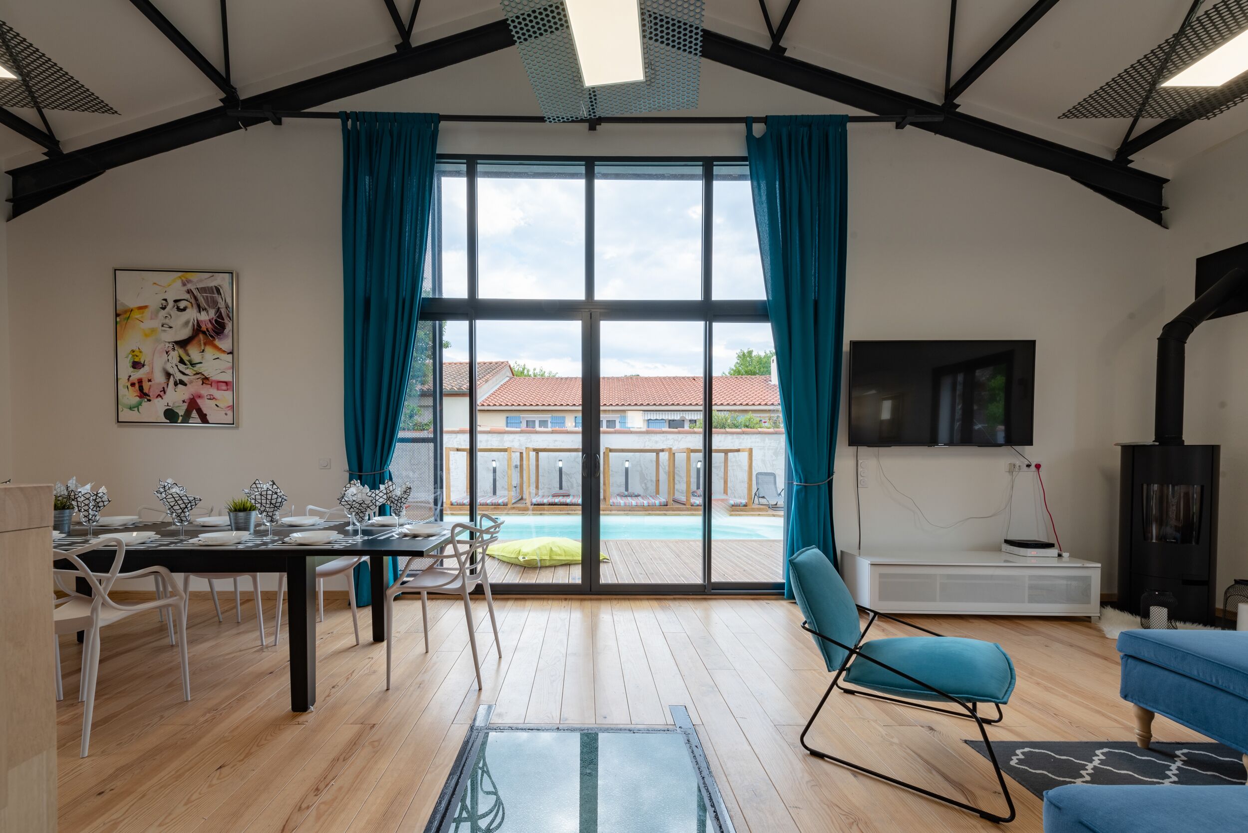 Een moderne woonkamer met een eethoek, grote ramen en uitzicht op een zwembad en gebouwen.