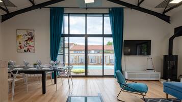 Een moderne woonkamer met een eethoek, grote ramen en uitzicht op een zwembad en gebouwen.