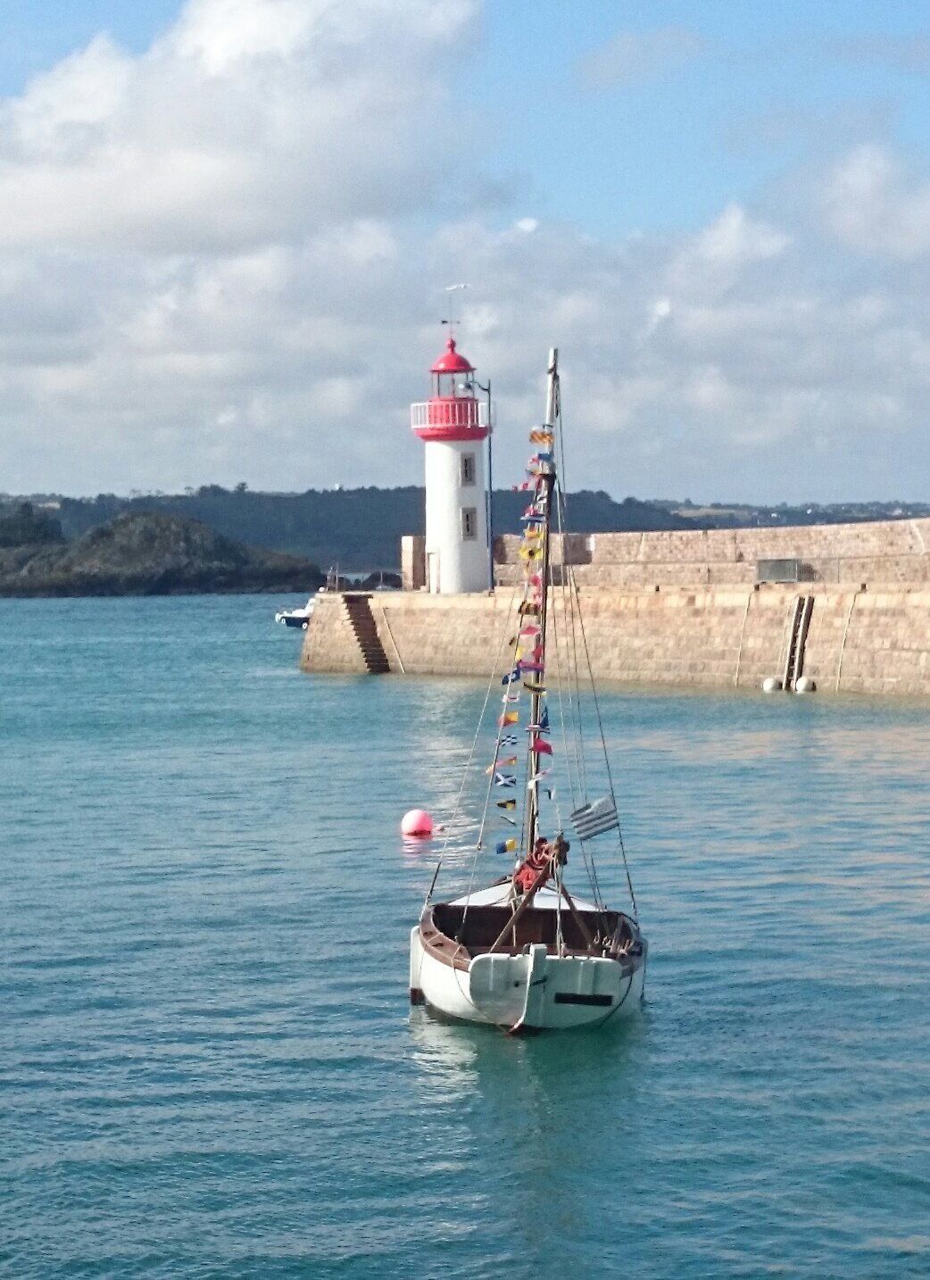 A boat with a tall mast and colorful flags, anchored near a lighthouse and a stone wall by the water.