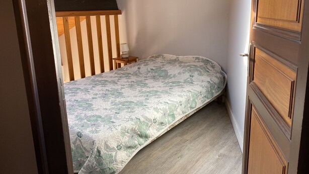 A bedroom with a wooden ceiling, a bed with a floral bedspread, a bedside table with a lamp, and a wooden door. - Bright and comfortable 3-room house in Saint-Palais, facing the sea (Saint-Palais-Sur-Mer)
