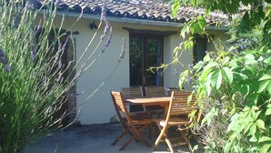 Een terras buiten met een houten tafel en stoelen, omgeven door planten en een huis met een pannendak.