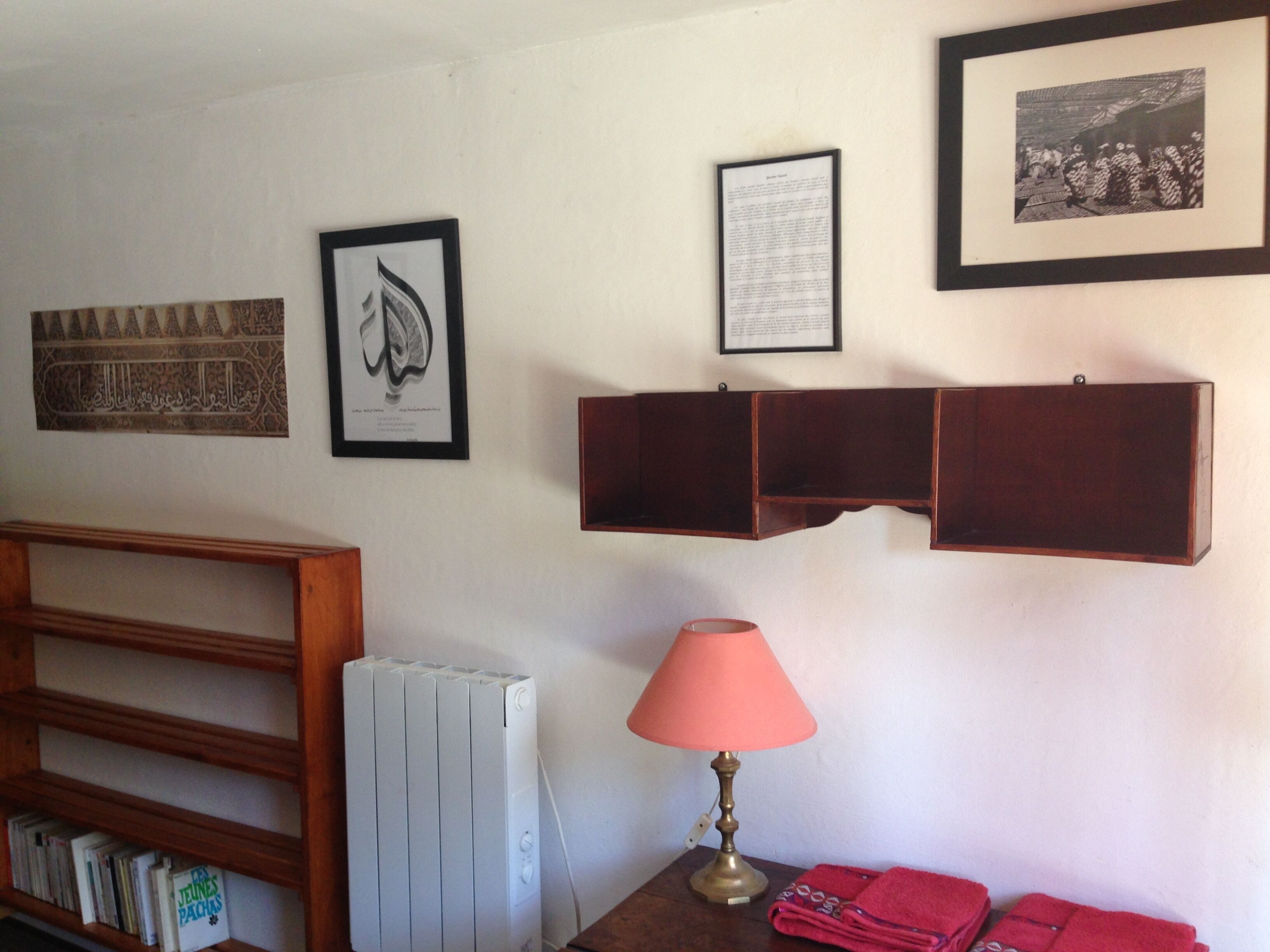 Una habitación con un radiador, una lámpara, una estantería con libros y objetos enmarcados en la pared.