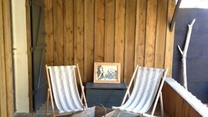 Eine Holzterrasse mit zwei gestreiften Liegestühlen, einem Metalltisch und einem gerahmten Bild an der Wand.