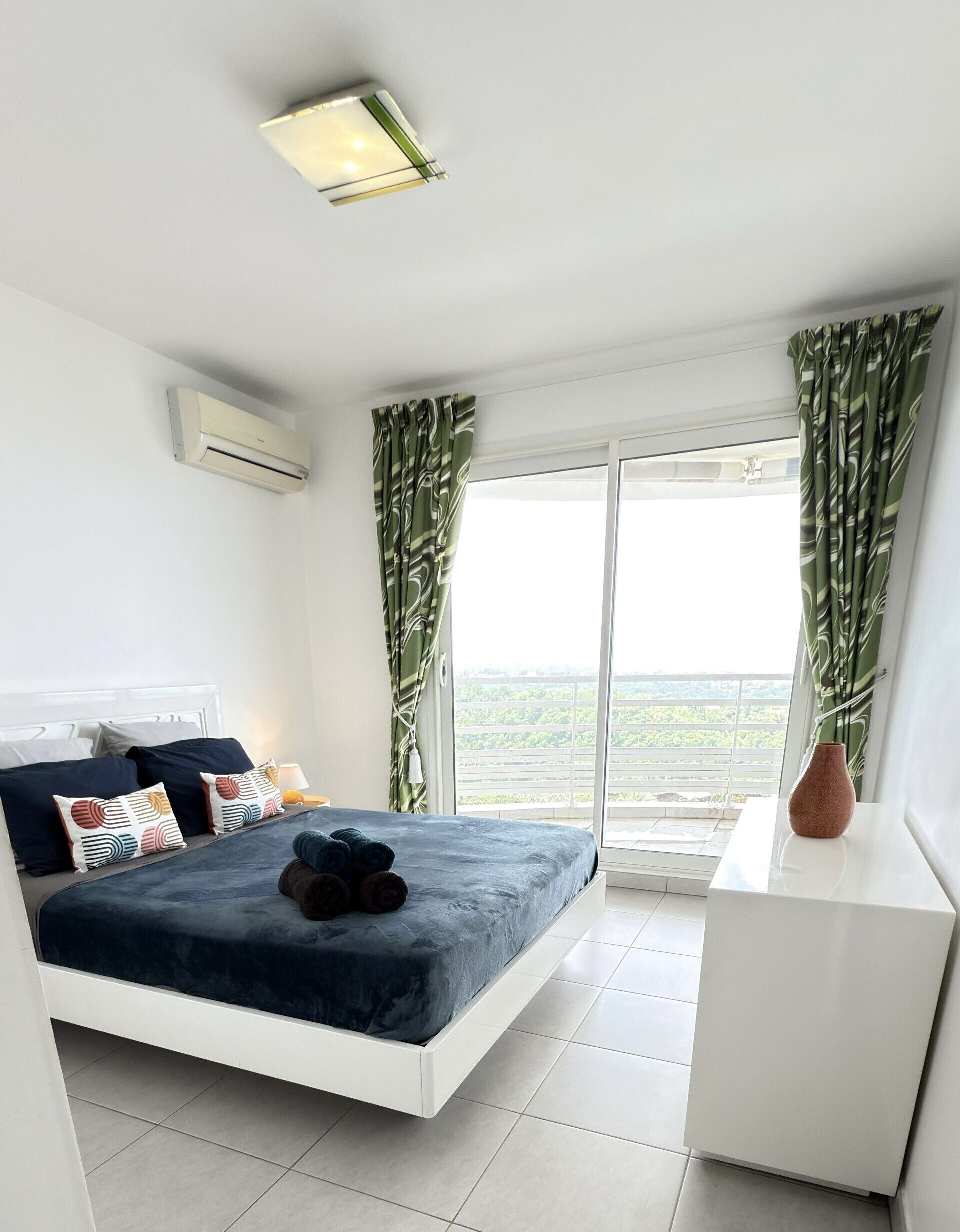 A modern bedroom with a bed, a nightstand, a vase, and a view of greenery through the window.