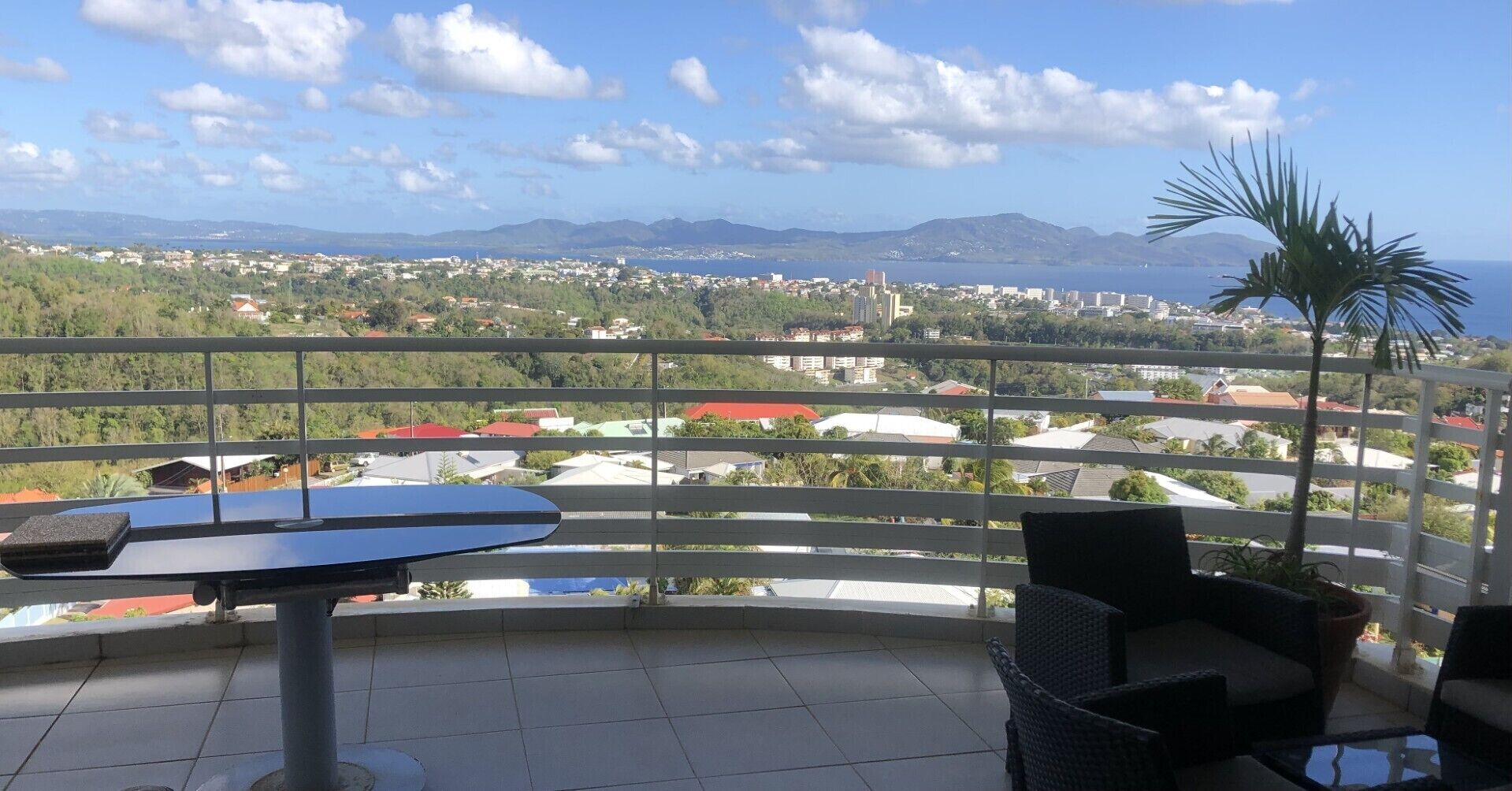 A balcony with a table and chairs, offering a view of a coastal town and mountains.