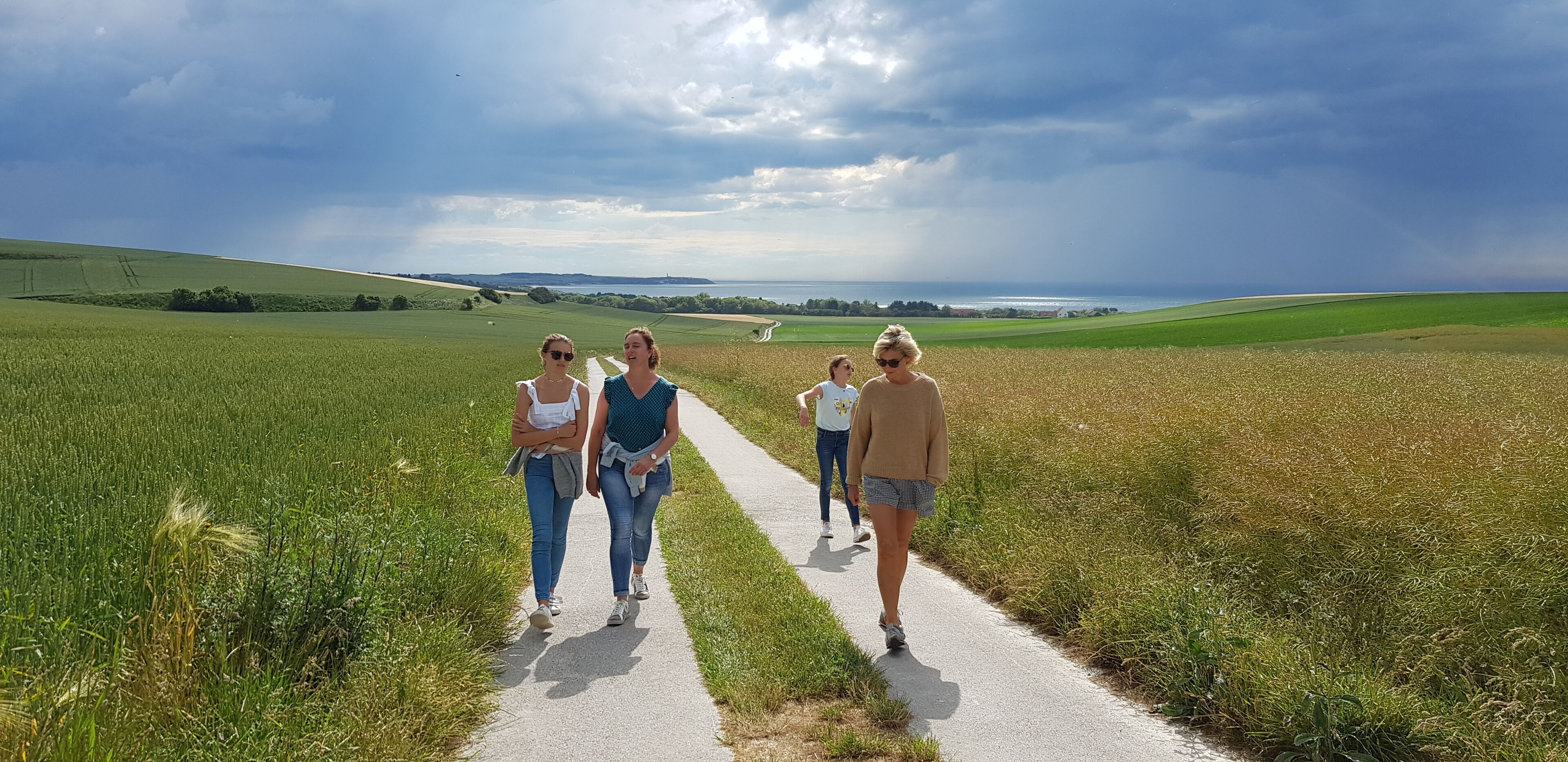 A paved path through a field with a view of the sea and hills in the distance.