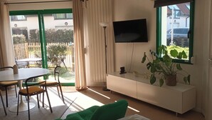 Ein Wohnzimmer mit Esstisch, Stühlen, einer Couch, einem an der Wand montierten Fernseher und einem Fenster mit Blick ins Freie.