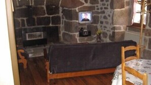 Huone, jonka seinät ovat kivestä, ja jossa on seinälle asennettu televisio, puinen sänky, kuvioidulla pöytäliinalla katettu pöytä ja tuoli.