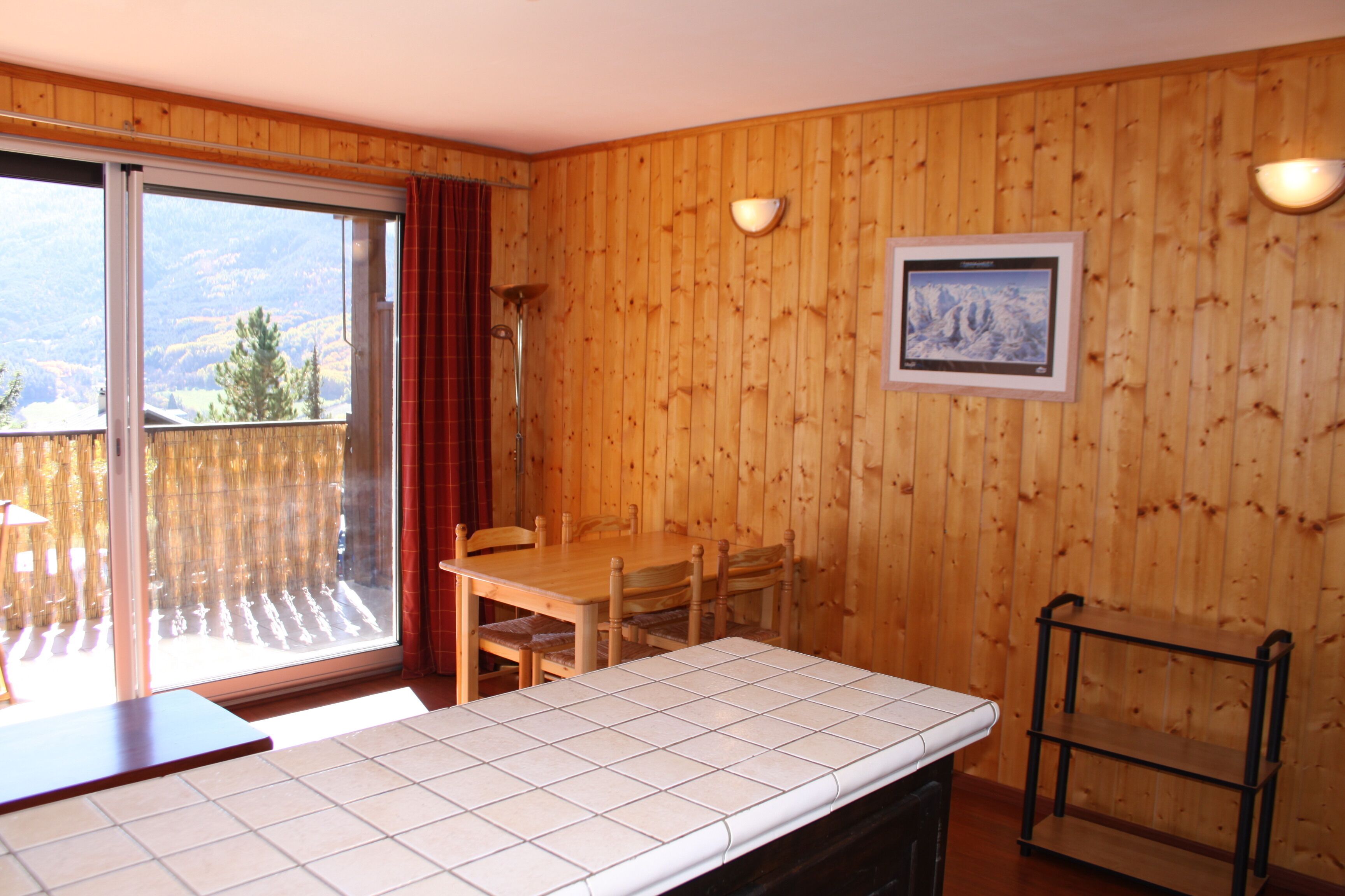 A wooden-paneled room with a dining table, chairs, a small shelf, and a sliding glass door leading to a balcony.