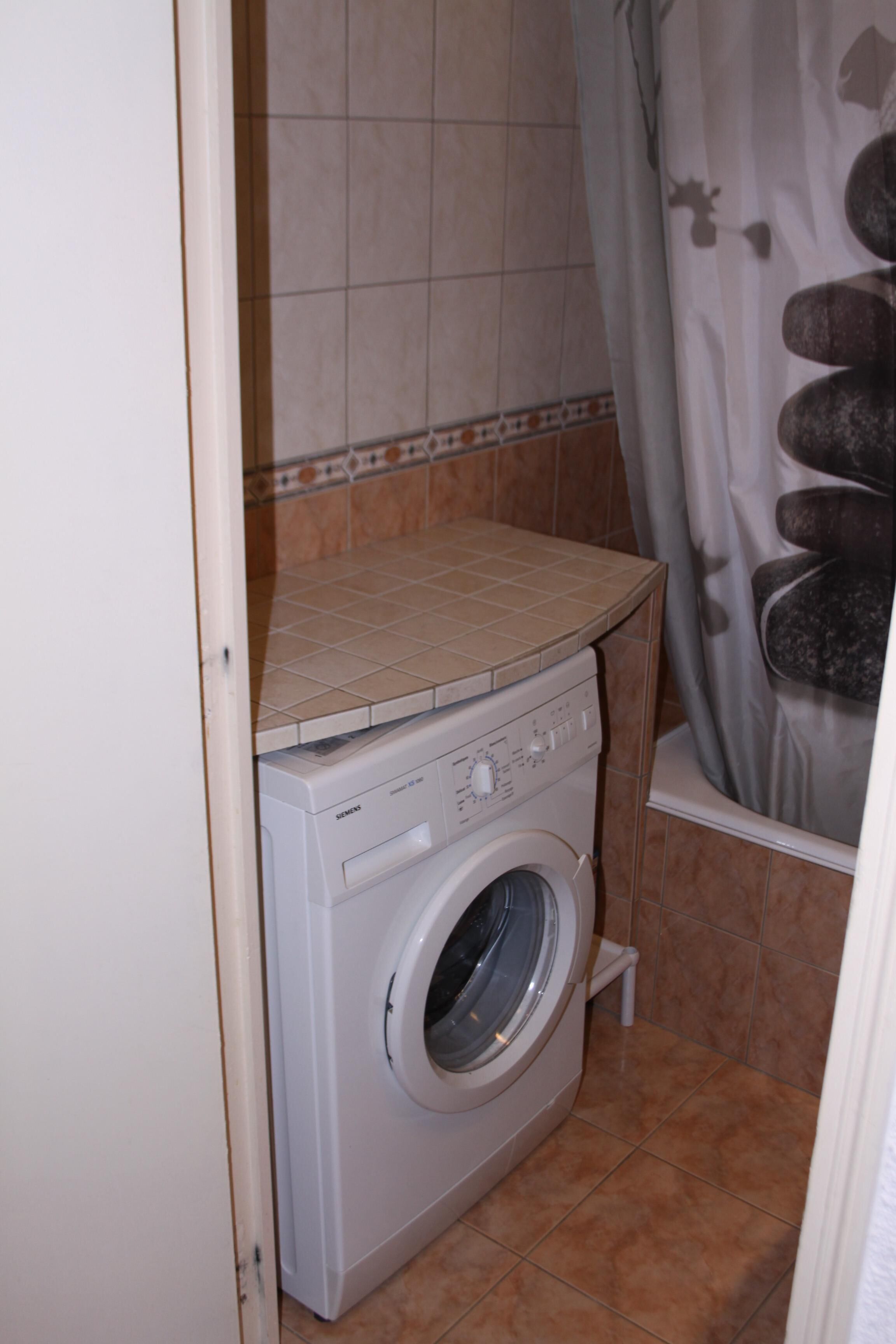 Een kleine wasruimte met een voorlader wasmachine, betegelde muren en een douchegordijn met een steenmotief.