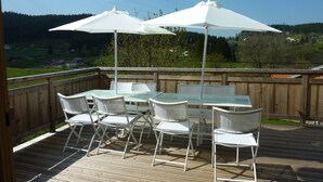 A wooden deck with a glass-top table and white chairs, surrounded by a wooden fence, overlooking a scenic landscape.