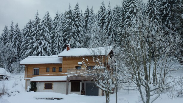 A wooden house with a snow-covered roof and a chimney, surrounded by snow-laden trees.