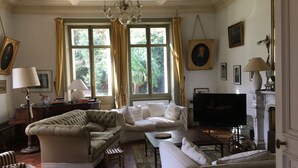 A spacious living room with a large sofa, a smaller sofa, a coffee table, and a television. There are two windows with curtains, a chandelier, and several framed pictures on the walls.
