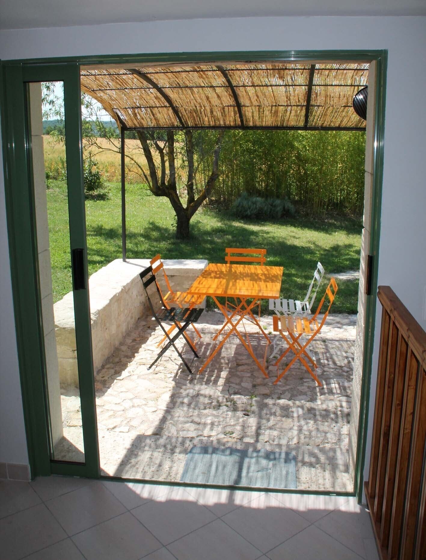 A patio with a table and chairs, a thatched roof, and a view of a tree and grass.
