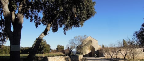 Un chemin de campagne mène à un bâtiment en pierre doté d’une entrée en arc de cercle, entouré d’arbres et d’un mur de pierre.