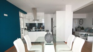 A modern dining area with a glass table and white chairs, a kitchen in the background, and a living area with a sofa and a clock on the wall. - Fully renovated apartment, in the Centre of Funchal. AMAZING view (Funchal)
