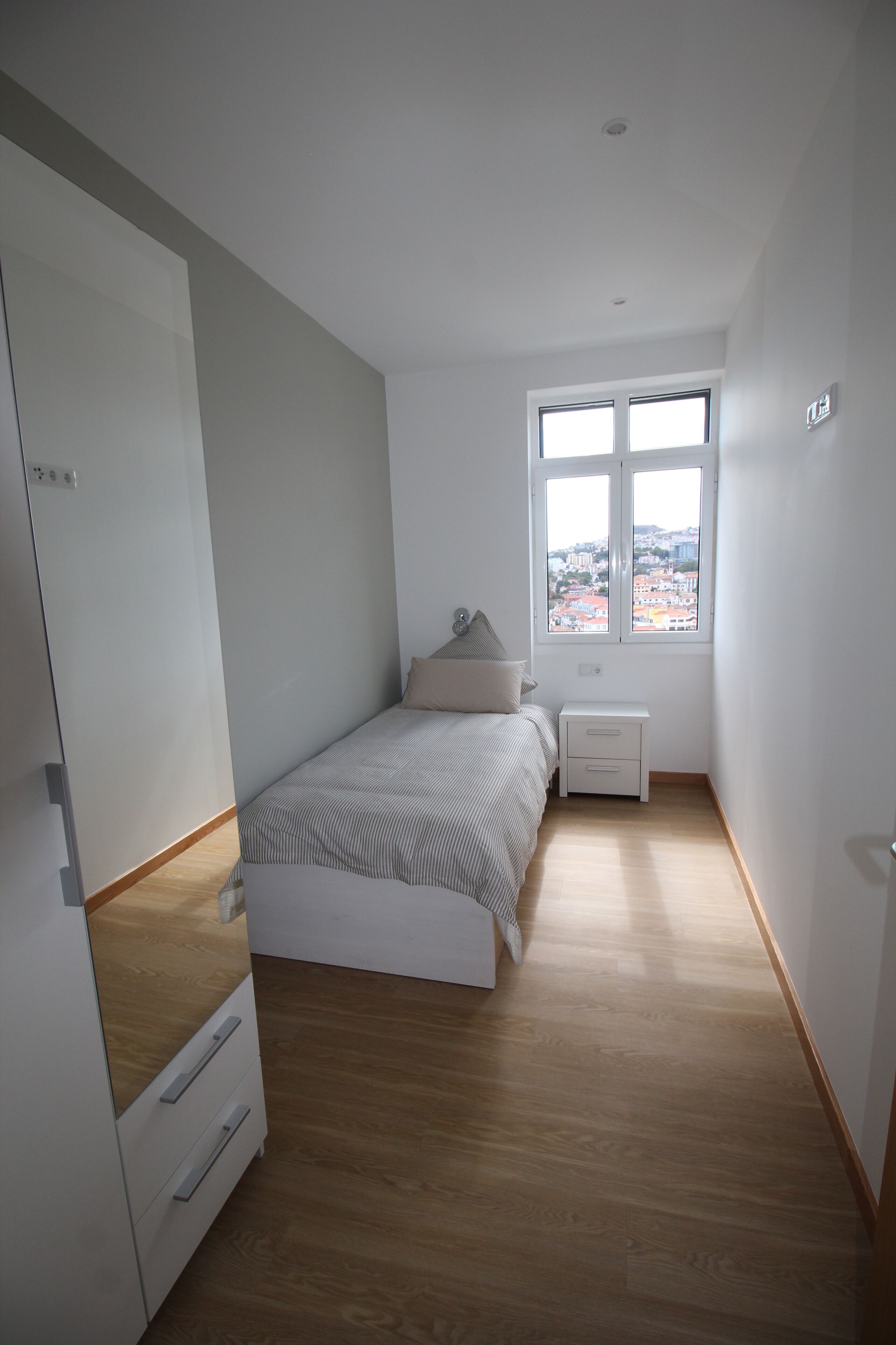 Une chambre à coucher comprenant un lit, une table de chevet, une commode et une fenêtre donnant sur la ville.