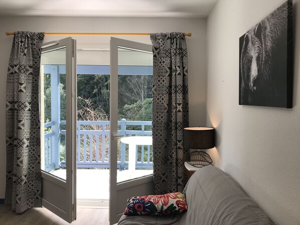 Una habitación con puerta de vidrio corrediza que da a un balcón exterior, un sofá con un cojín colorido, una lámpara de pared y un cuadro enmarcado.