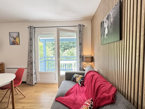 A living room with a grey sofa, a red blanket, a patterned pillow, a table with a red chair, and a view of a balcony with blue railings.