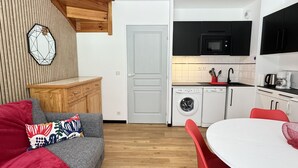A compact living space with a grey sofa, red cushions, a wooden accent wall, a kitchen with a microwave, washing machine, and a dining area with a table and chairs.