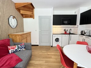 Un espacio habitable compacto con un sofá gris, cojines rojos, una pared de acento de madera, una cocina con microondas y lavadora, y una zona de comedor con mesa y sillas.