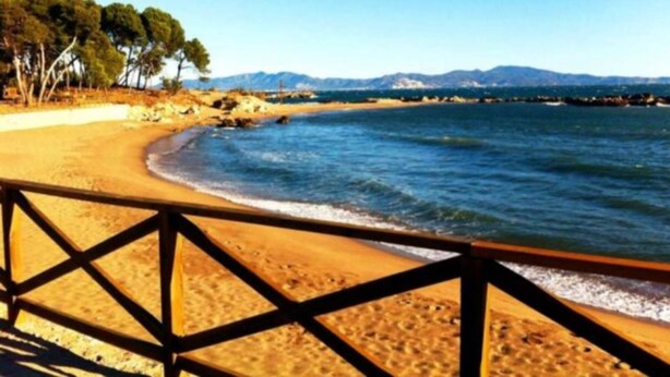 Une plage avec une clôture en bois, un rivage sablonneux et une vue dégagée sur la mer et les montagnes lointaines.