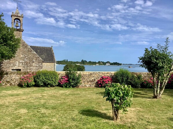 A stone building with a bell tower, a grassy area with flowering bushes, and a view of a body of water with boats. - Charming villa in Saint-Philibert (close to Carnac) directly by the sea. 3*rated (Saint-Philibert)