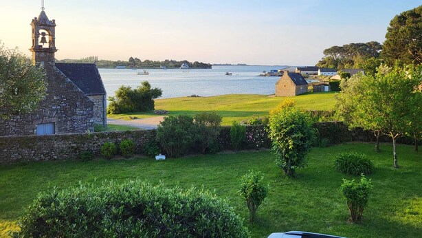 En byggnad av sten med ett klocktorn, en gräsyta och utsikt över vattnet med båtar.