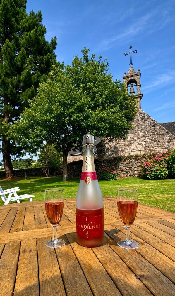 A bottle of FREIXENET wine and two glasses on a wooden table with a stone church and greenery in the background. - Charming villa in Saint-Philibert (close to Carnac) directly by the sea. 3*rated (Saint-Philibert)
