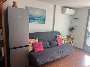 A living room with a grey sofa, a painting of a seascape, a white air conditioning unit, and a small table with a vase.