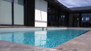 A modern pool with clear blue water, surrounded by a stone deck and a building with large glass windows. - Magnificent villa with heated pool close to beaches (Arzon)