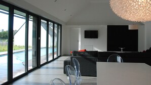 A modern living room with a large glass door leading to a pool, a black sofa, and a mounted TV. - Magnificent villa with heated pool close to beaches (Arzon)