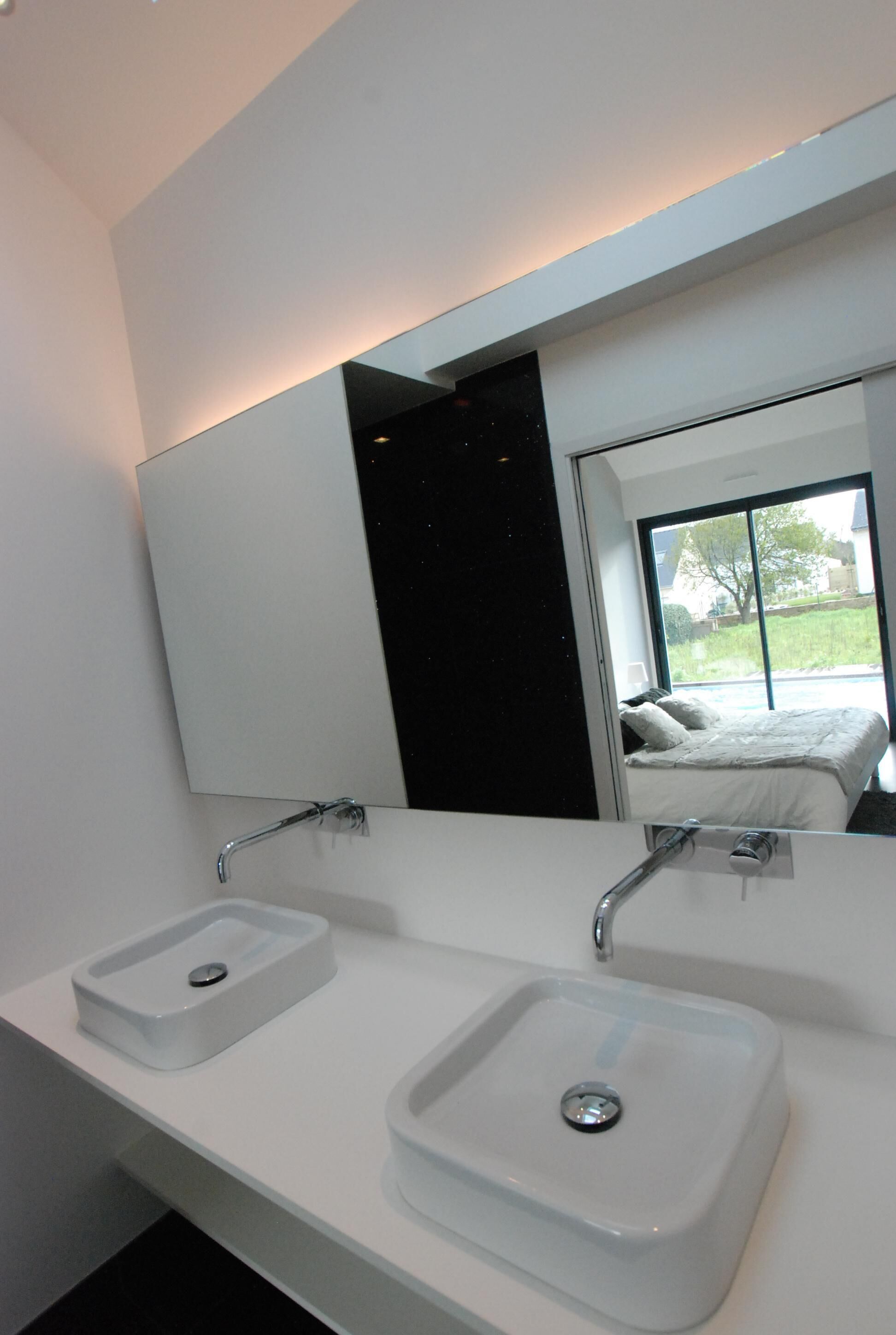 A modern bathroom with two white sinks, a large mirror, and a view of a bedroom with a bed and window.