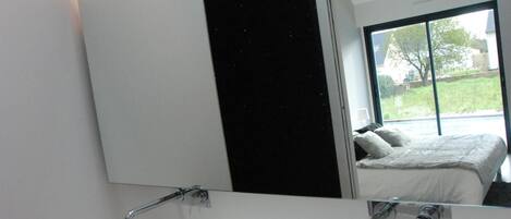 A modern bathroom with two white sinks, a large mirror, and a view of a bedroom with a bed and window.