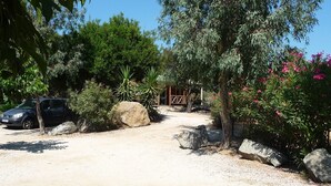 A gravel driveway leading to a wooden structure, surrounded by various plants and a parked car. - Algajola - New wooden air-conditioned chalet for 4/5, 1 km away from the beach (Algajola)