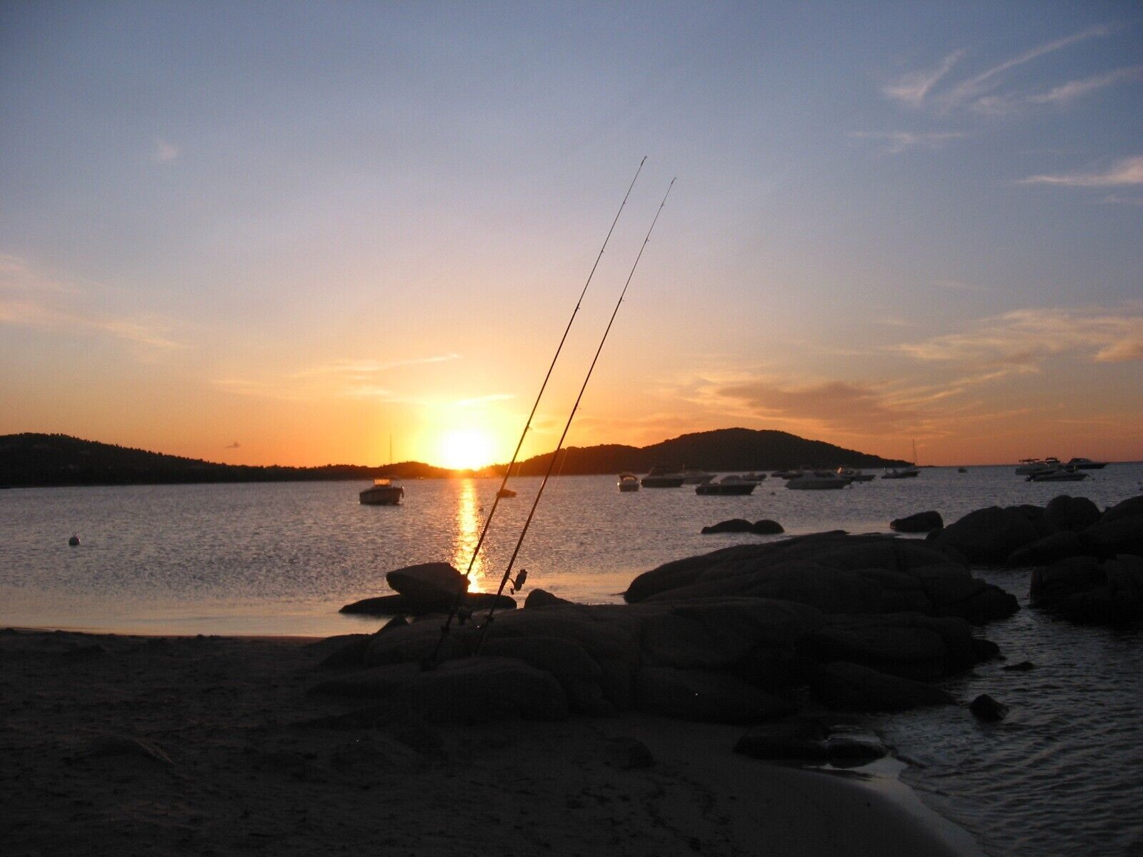 A sunset over a calm body of water with boats and fishing rods on the shore.