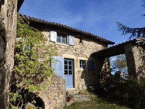 Un bâtiment en pierre avec une porte et des volets bleus, entouré de verdure.