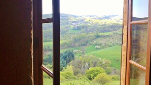 Blick durch ein Fenster auf eine ländliche Landschaft mit sanften Hügeln, Bäumen und einem Gebäude in der Ferne.