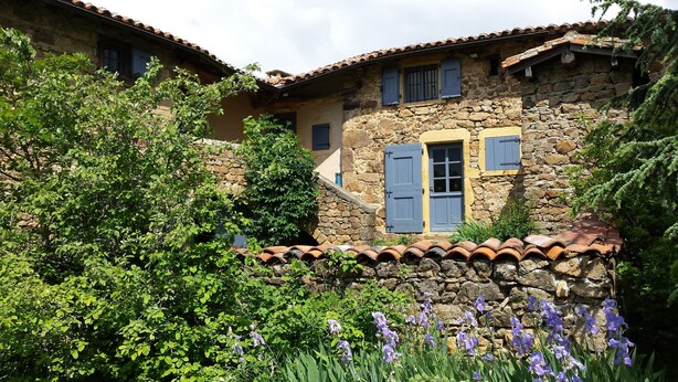 Ein Gebäude aus Stein mit einem Ziegeldach, blauen Fensterläden und einem Garten mit violetten Blumen.